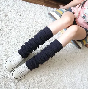 Close-up of black knitted slouch leg warmers worn with white sneakers on a rug.