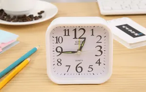 Close-up of a minimalist white square alarm clock on a wooden desk with pencils and coffee.