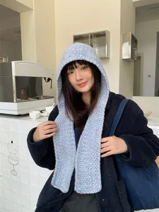 Model wearing light blue knitted hooded scarf in a cozy cafe setting for winter warmth.