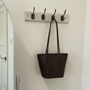 Donkerbruine gevlochten leren tote bag hangend aan een minimalistische wandhaak in de kamer.