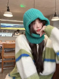 Close-up of a green knitted balaclava hood worn by a model in a cozy cafe setting.