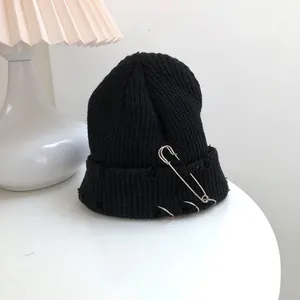 Close-up of a distressed black knit beanie featuring silver metal rings and a large safety pin detail