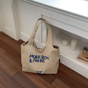 Bolso tote de lona beige casual con texto 'MORE RICH & MORE' bordado en azul y logo