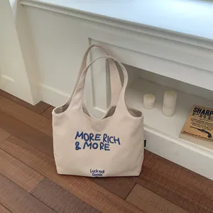 Detalle de un bolso tote de lona color crema con eslogan bordado en azul More Rich