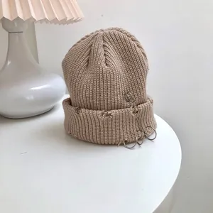 Detail of a beige ribbed knit beanie with silver safety pin and ring accents.