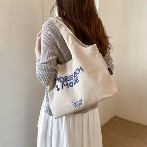 Bolso tote de lona beige con letras azules bordadas al hombro de una modelo