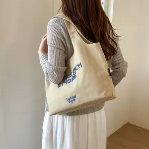 Primer plano de un bolso tote de lona beige con bordado azul llevado por una modelo.