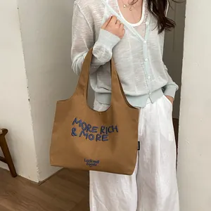 Bolso tote grande de lona marrón informal con bordado 'MORE RICH & MORE' sostenido por modelo.
