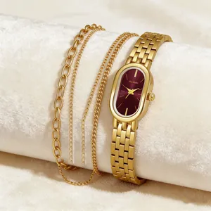 Nahaufnahme einer bordeauxroten ovalen Uhr und goldenen Armbändern auf weißem Plüsch.