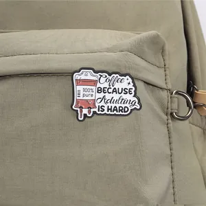 Close-up of "Coffee Because Adulting Is Hard" IV bag enamel pin on a green backpack.