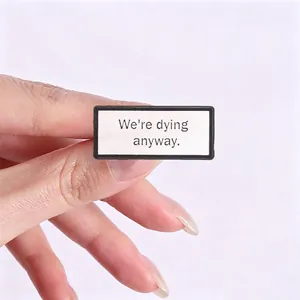 Close-up of a minimalist square metal badge with We're dying anyway english quote