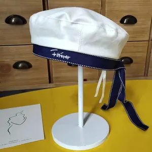 Detail view of white beret with navy embroidered ribbon and long bow tie decoration