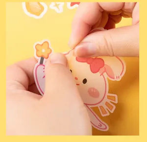 Close-up of hands peeling a cute waterproof cartoon PVC sticker from a yellow surface