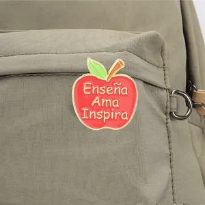 Close-up of red apple enamel pin with Spanish Teach Love Inspire text on backpack