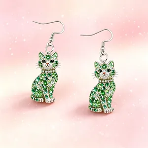 Close-up of crystal-encrusted green cat drop earrings on a sparkling pink background.
