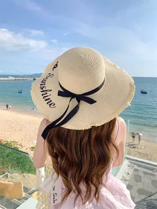Ψάθινο καπέλο με φαρδύ γείσο, κέντημα Sunshine και μαύρο φιόγκο σε μια ηλιόλουστη παραλία.