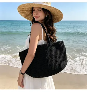 Model im weißen Kleid mit schwarzer Strohtasche am Strand unter blauem Himmel.