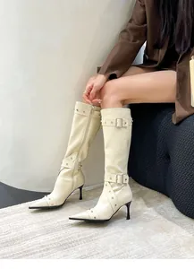 Detail view of cream pointed-toe knee-high boots featuring buckle and stud accents