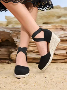Elegantes sandalias de esparto negras con cuña y tiras cruzadas sobre fondo de arena