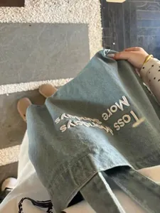 Detailed view of embroidered 'Less More' text on a green retro canvas tote bag for school.