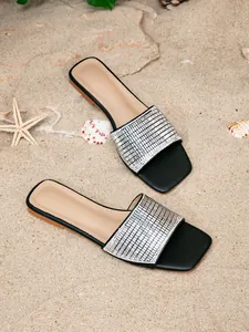 Detail of black flat sandals with sparkling rhinestone strap on beach sand.