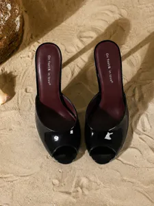 Κορυφαία όψη μαύρα λουστρίνι stiletto mules σε φόντο με ψιλή άμμο παραλίας
