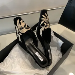 Detalle de mules de terciopelo negro con elegantes bordados florales blancos en caja.