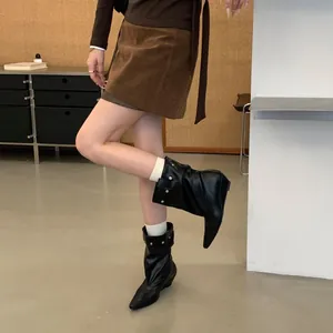 Close-up of black pointed-toe slouchy knight boots with decorative belt buckle and wedge heel.