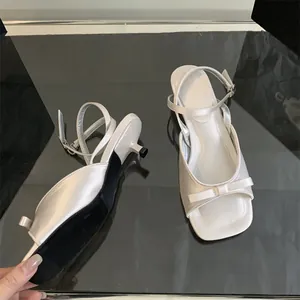 Detail view of white satin kitten heel sandals with bow and ankle strap on glass surface.