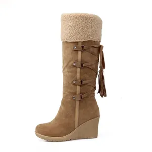 Vista lateral de botas altas de ante marrón con puño de sherpa, borlas y tacón de cuña.