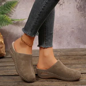 Detail view of women's taupe wedge mule slippers with white stitching on rustic wood floor