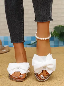 White flat sandals for women featuring large bow decor and rhinestone trim on a beach setting