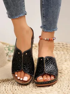 Close-up detail of black croc-pattern wedge slippers with gold studs on a straw mat