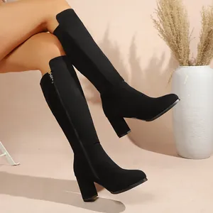 Elegant black suede knee-high boots with chunky heels on a model in a stylish interior.