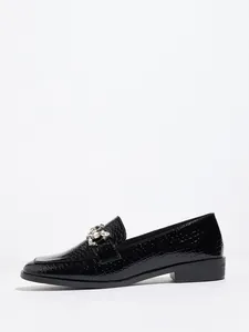 Detalle de mocasines de charol negro con grabado de cocodrilo y hebilla de cristal.