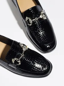 Primer plano de mocasines negros brillantes con efecto cocodrilo y detalle de pedrería
