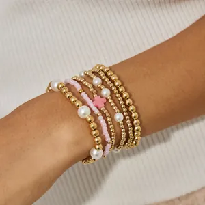 Detail van een stapel gouden kralenarmbanden met parels en een roze kruisbedeltje
