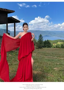 Elegant red one-shoulder pleated maxi dress with side slit posing in a scenic mountain landscape