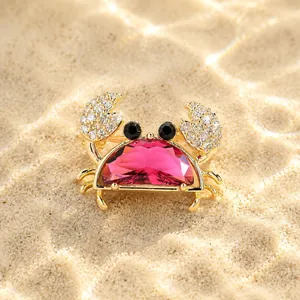 Detailed macro view of a gold-toned crab brooch with a vibrant pink crystal body on sand