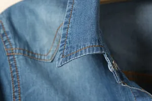 Détail du col d'une chemise en jean bleu avec surpiqûres orange et un bouton métallique.