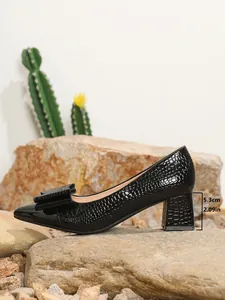 Close-up of black crocodile-textured pump with bow and 5.3cm block heel on stone.