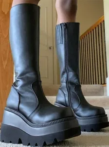 Side view of black platform knee-high boots with thick wedge lug soles on carpeted stairs