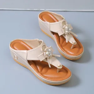 Detalle de chanclas beige con cuña, adorno floral de perlas y detalle de letra dorada.