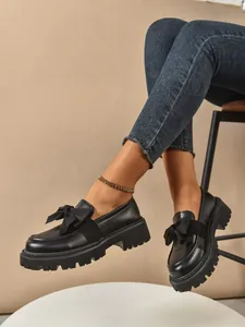 Close-up of black platform loafers with a decorative bow and thick lug sole.
