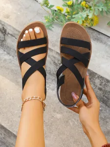 Stylish women's flat sandals with black elastic criss-cross straps shown on stone steps.