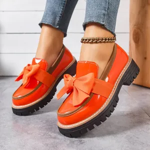 Close-up of vibrant orange patent leather loafers featuring a large bow and thick sole