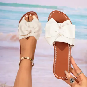 Detail view of women's flat slippers with a large white bow on a beach background