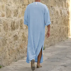 Back view of a man walking in a long blue cotton linen robe against a stone wall.