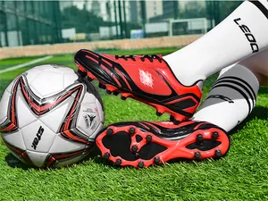 Detailansicht der rot-schwarzen AG-Fußballschuhe auf grünem Kunstrasen mit einem Ball.