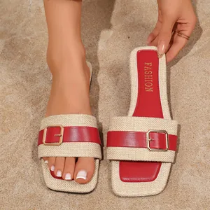 Nahaufnahme flacher Strandsandalen mit roter Schnalle und beigem Webmuster auf Sand.